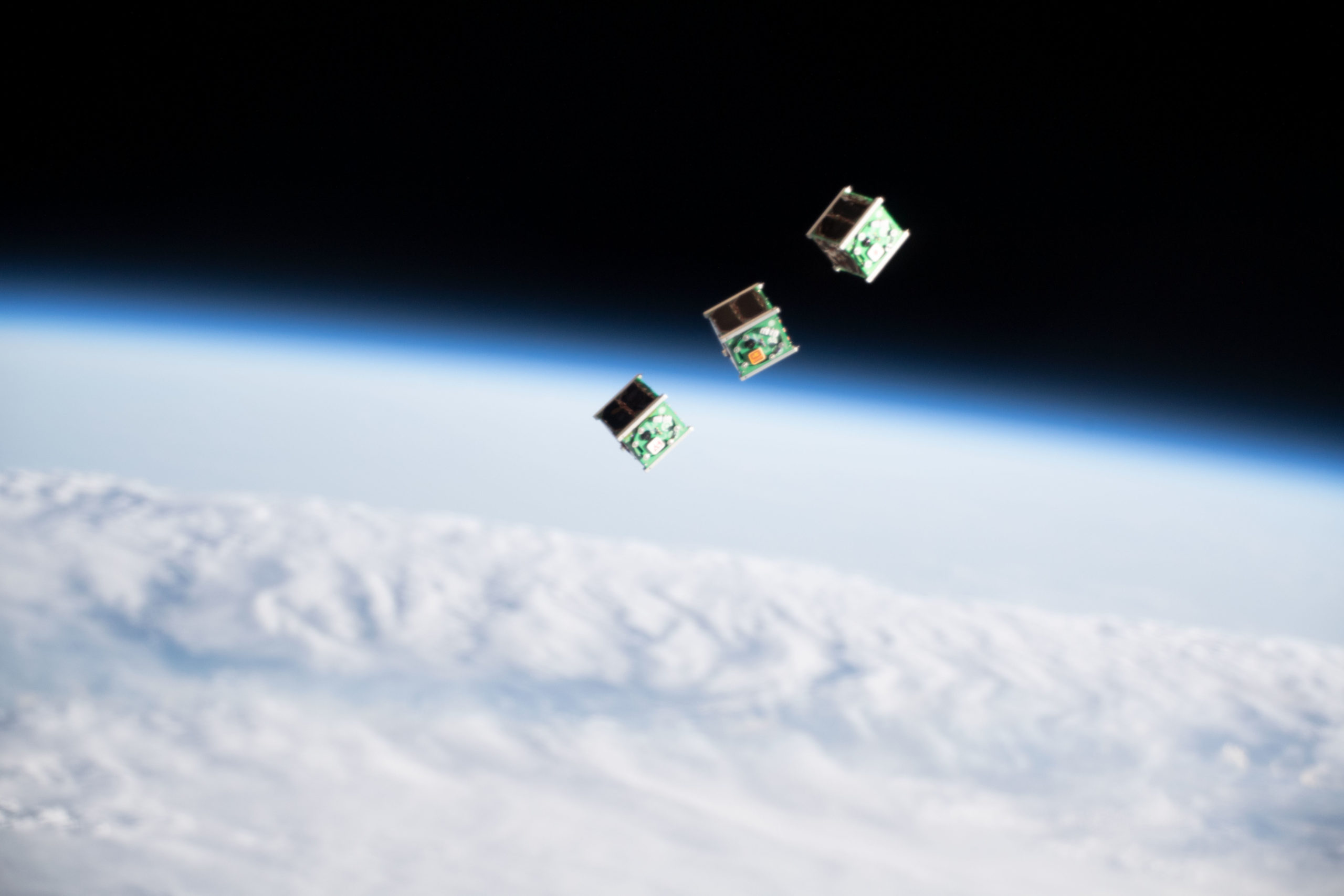 Three cubesats in space with cloud-covered Earth and dark space as a backdrop. Photo from the International Space Station by NASA.